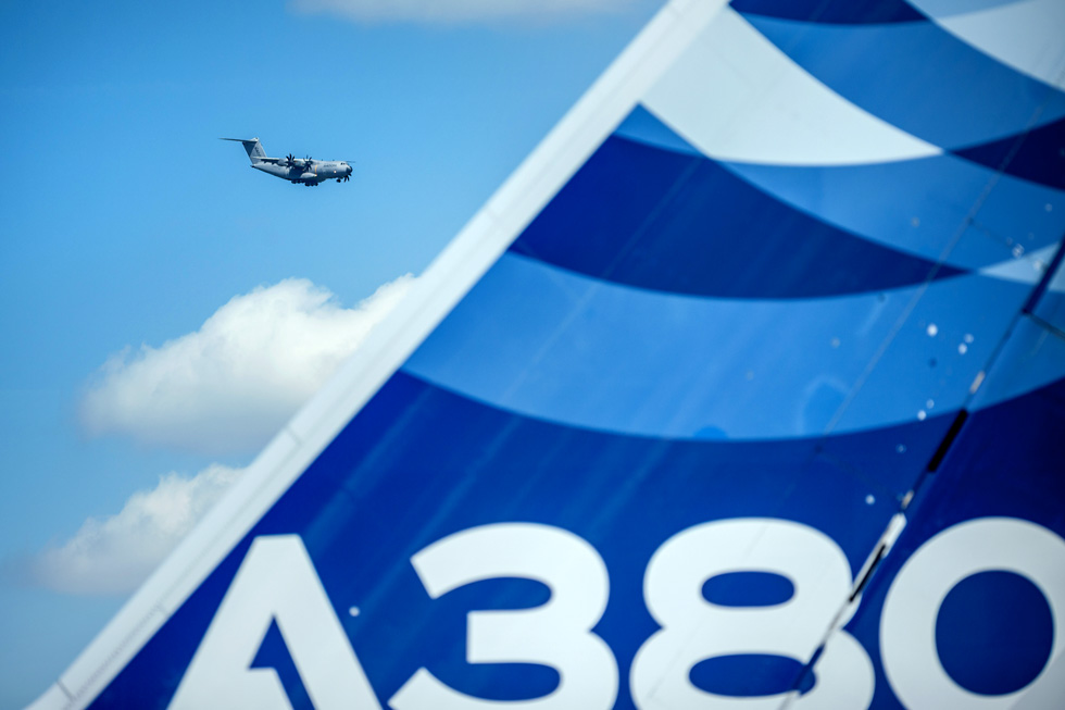Im Fokus oben links ein fliegendes Flugzeug vor blauem Himmel. in der rechten Bildhälfte ist unscharf eine Heckflosse des A380 erkennbar.