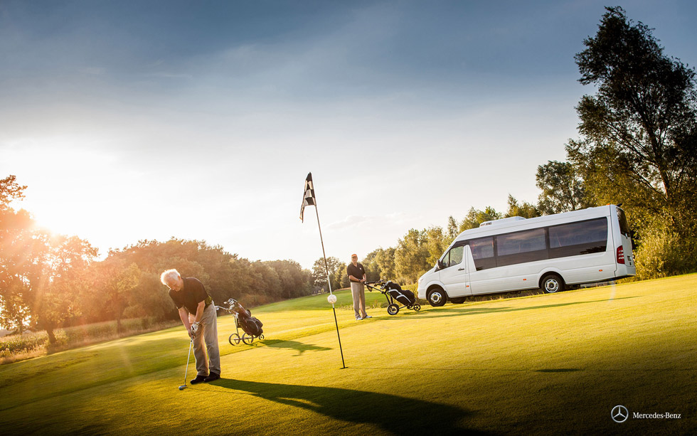 Auf einem Golfplatz schlägt ein Mann einen Ball. Im Hintergrund steht ein weiterer Golfer mit Golftasche neben einem Minibus von Mercedes Benz.