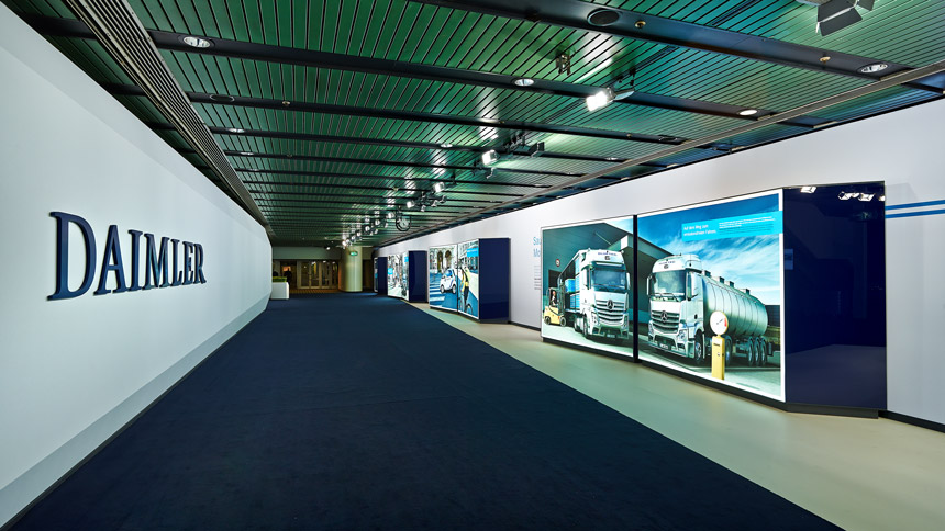 Ein Gang innerhalb eines Gebäudes mit blauem Teppich. Auf der linken Wand steht DAIMLER und auf der rechten Seite stehen Boxen mit Fotos LKWs.