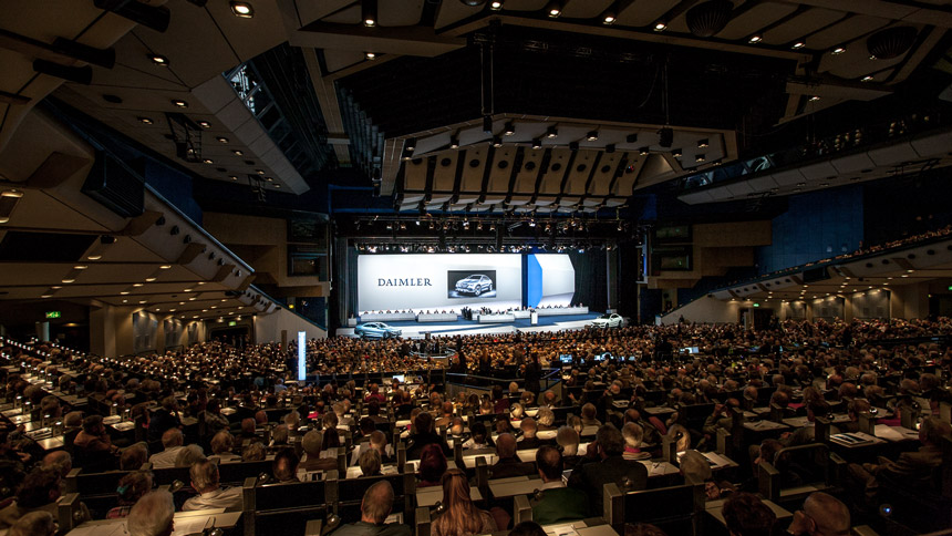 Aussicht vom Publikum auf eine Bühne, auf der Autos stehen und Menschen sitzen. Im Hintergrund ist ein Foto eines Wagens und der Schriftzug Daimler zu sehen.