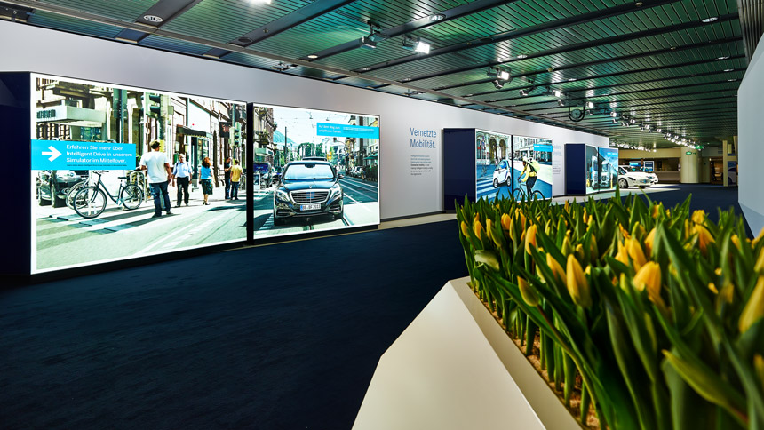 Im Innenbereich ein Gang mit blauen Teppich, gelbe Tulpen als Dekoration auf der rechten Seite. Auf der Linken Seite stehen menschgroße Boxen mit Displays, die Fotos von verschiedenen Fahrzeugen zeigen.