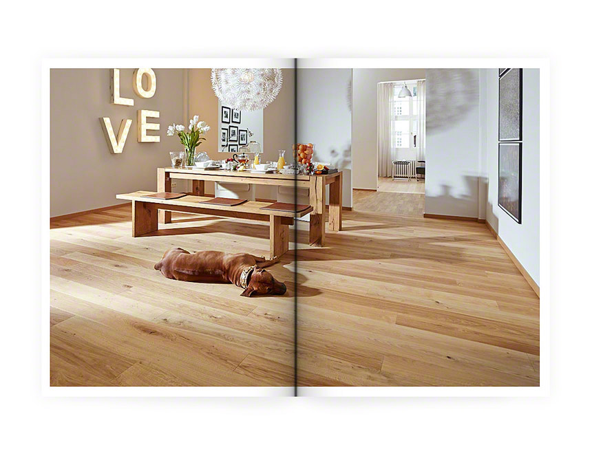 Eine aufgeschlagene Zeitschrift mit einem ganzseitigen Foto mit Blick in ein Esszimmer. Ein heller Holzfusboden auf dem ein ähnlich brauner Tisch steht, der fertig gedeckt fürs Frühstück ist. Vor dem Tisch liegt ein dunkelbrauner Hund.
