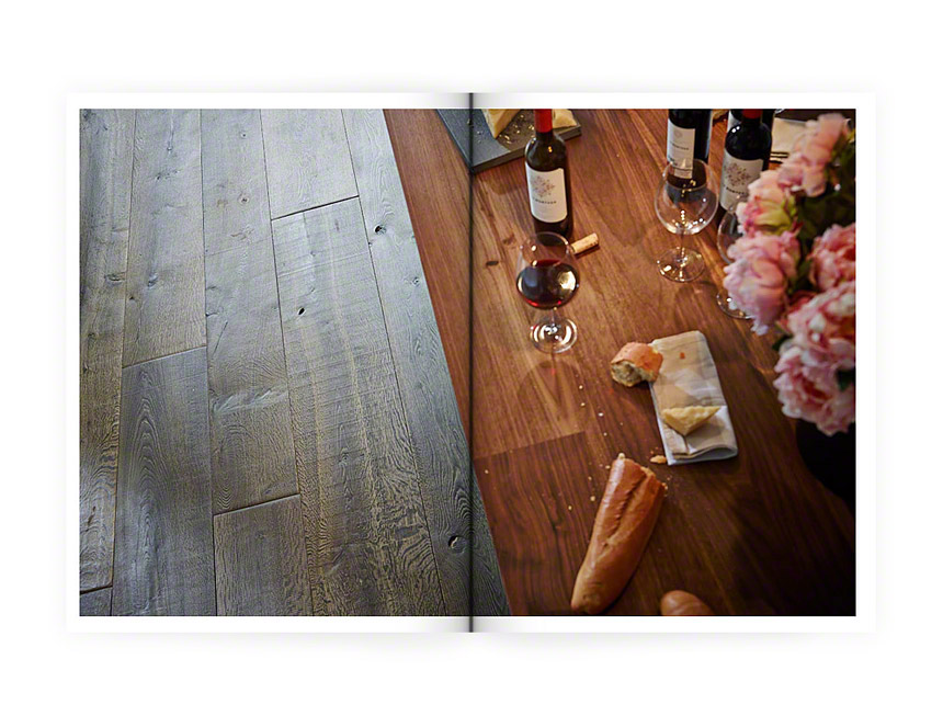 Eine aufgeschlagene Zeitschrift mit Blick auf einen Tisch aus der Vogelperspektive. Auf dem Tisch sind Weingläser, Blumen und ein Baguette zu sehen. Neben dem Tisch sieht man durch die Perspektive den Holzboden.