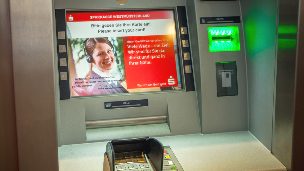 Blick auf einen Geldautomaten. Der Bildschirm zeigt ein Werbefoto mit Text.