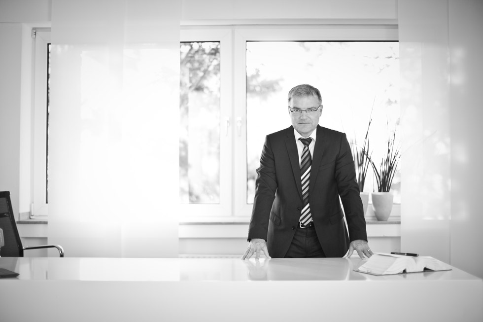 Portraitaufnahme in Schwarz Weiss von einem Mann im Anzug, der an einem weissen Schreibtisch steht und in die Kamera schaut.