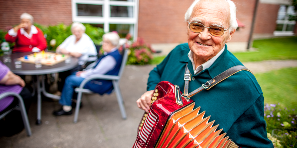 Ein älterer Herr spiel draußen ein Instrument. Im Hintergrund sitzt eine Gruppe am Tisch