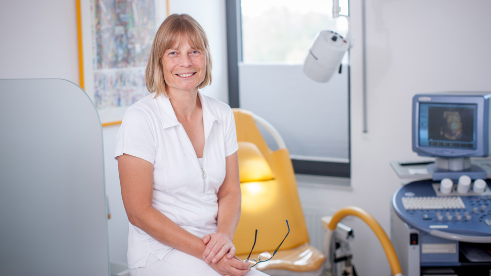 Eine Dame sitzt in einem Behandlungsraum. Im Hintergrund ist ein GynäkologenStuhl und Ultraschall gerät zu erkennen.