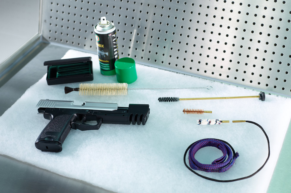 Auf weisser Watte liegt eine Pistole, Reinigungsbürsten, eine Spraydose und eine Schachtel.