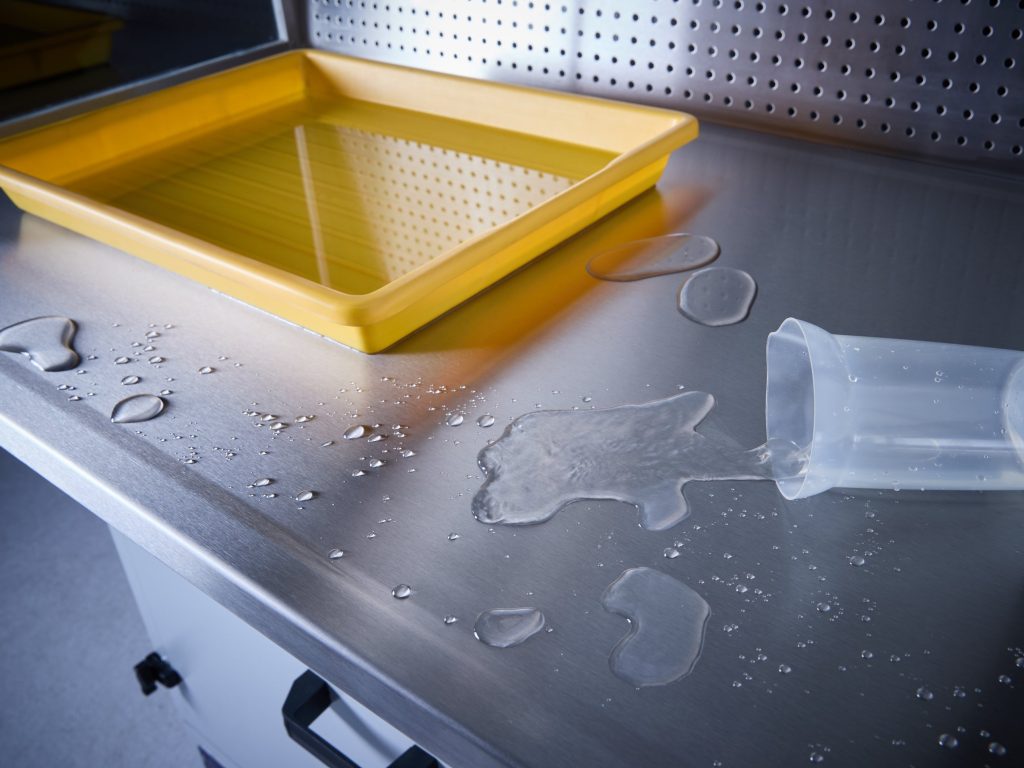 Wassertropfen und ein umgekippter Messbecher auf der Edelstahl-Arbeitsfläche eines Absaugtisches, im Hintergrund eine gelbe Schale